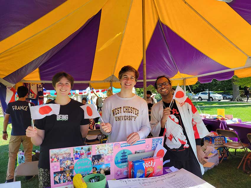 Three students standing at a table under an event tent. They are holding small flags for Japan.