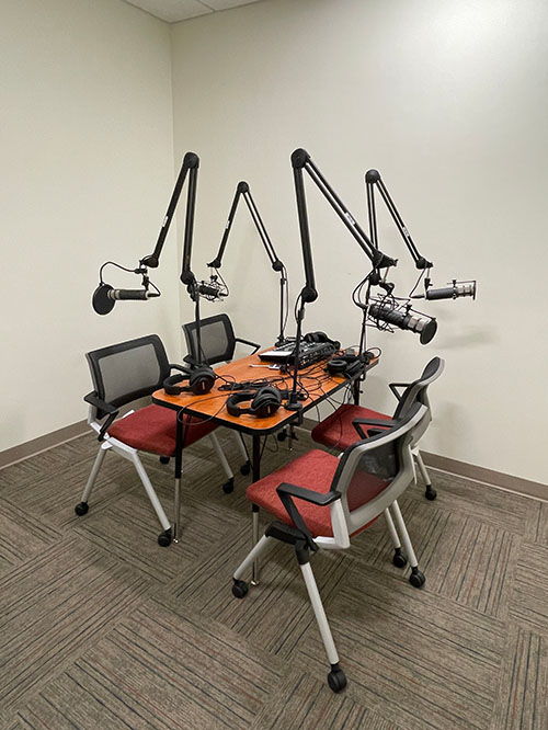 A photo of the podcast setup in the audio recording studio. Pictured is a Rodecaster Pro Mixer along with 4 microphones with boom arms attached to a rectangular square table.