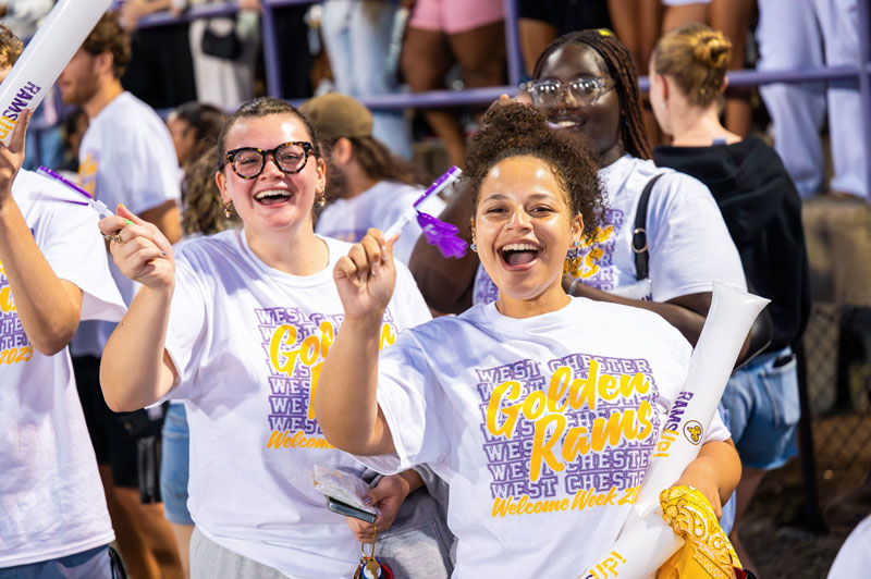 Two students picured wearing golden rams tshirts and waving wcu clapper toy at a football game