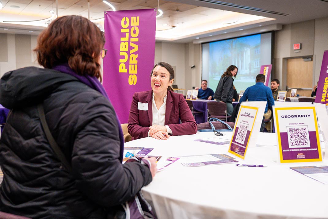 Two people sitting at a table in front of a Public Service sign for Graduate School Open House.