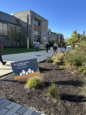 Campus walkway beside a modern stone building with students walking in the distance. In the foreground, a small yard sign with cartoon penguins displays a short message.
