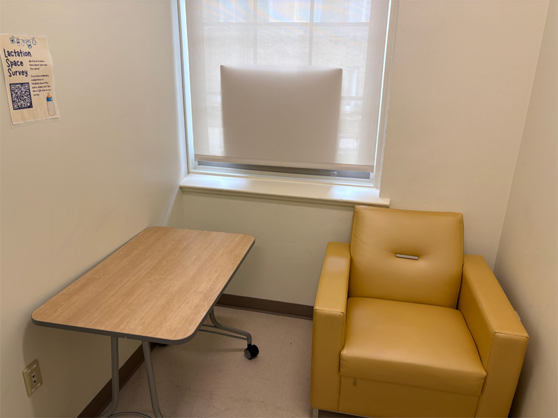 From door entry, a small wooden table sits next to an armchair which sits in front of a window with protective covering.  