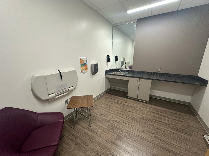 From door entry, there is an armchair by the front and left walls, with a side table. On the left wall, there is a changing table. On the back wall, a sink and long counter with a storage cabinet underneath. 