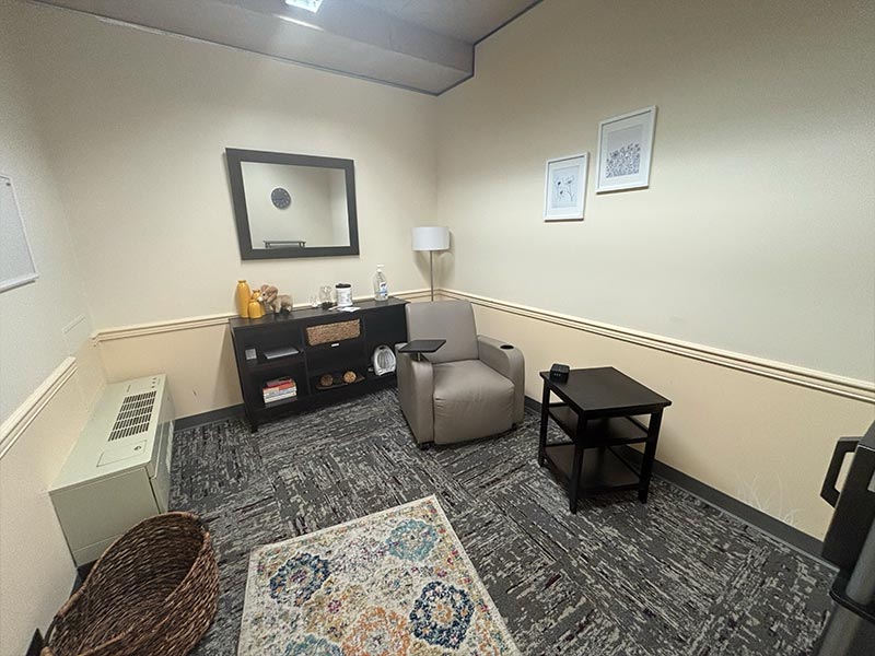 From door entry, the room is inviting with a rug, decor on the back wall including storage and a table top, and an armchair close to the right wall. Next to the armchair is a small side table. On the front wall, a microwave. 