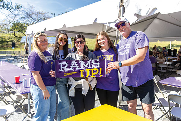 A group of people smile and pose for a picture while wearing their WCU gear. They are holding a sign that says Rams Up!