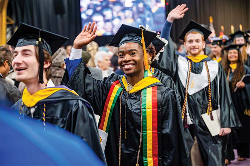 A group of graduates in caps and gowns walk in a ceremony, smiling and waving to the audience.