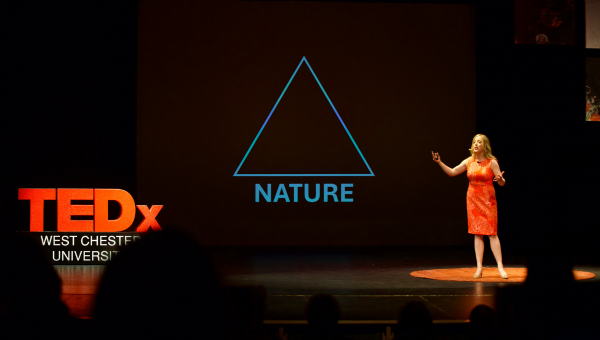 A female speaker at WCU's TEDx event. She is on a large, dark stage with the words "TEDX West Chester Univeristy" in lights on the left side of the photo. 
