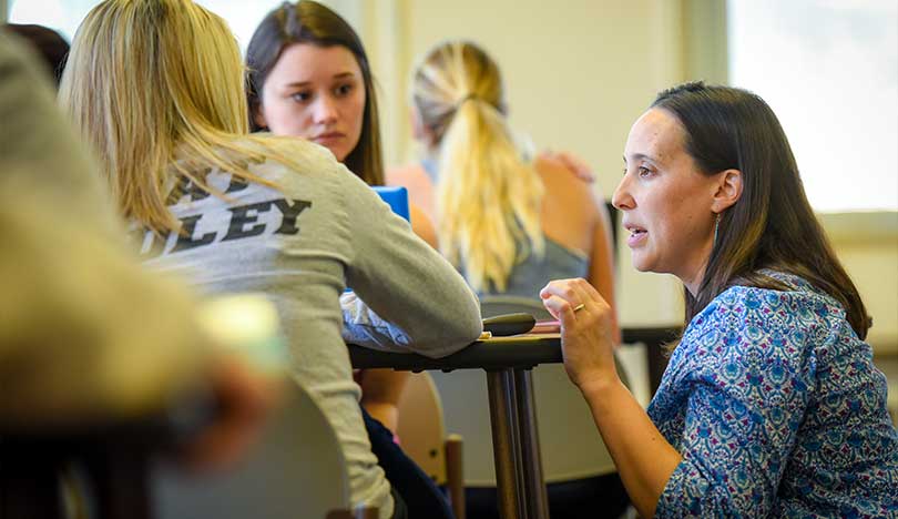 An instructor kneels down to students sitting in the classroom at a  table