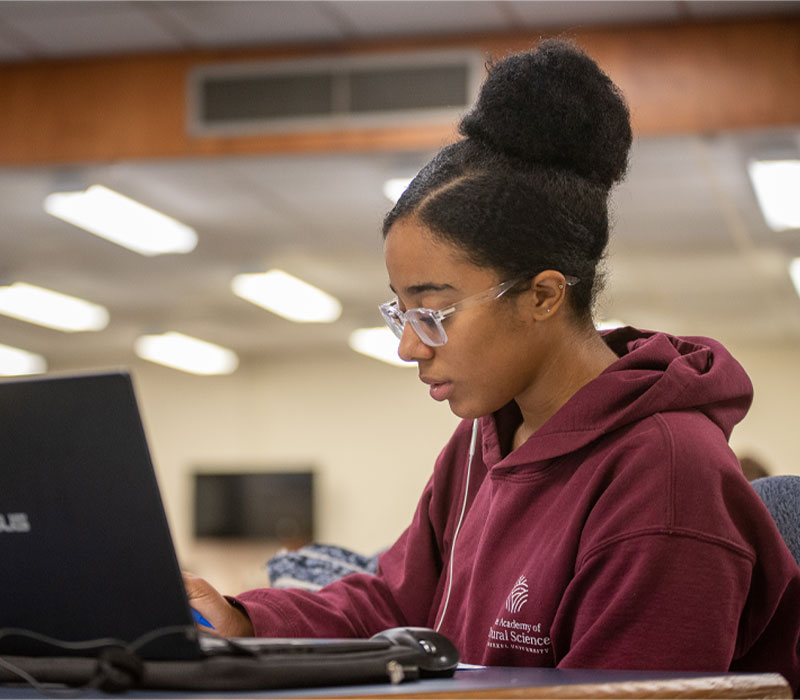 A student wearing glasses and a red sweatshirt is working on her laptop