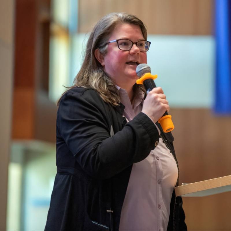 individual with brown hair, black jacket and purple shirt speaking into a microphone