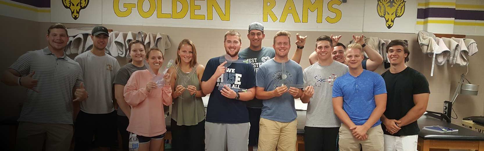 Twelve young adults pose in a school training room under a “Golden Rams” sign, several holding clear plastic items.
