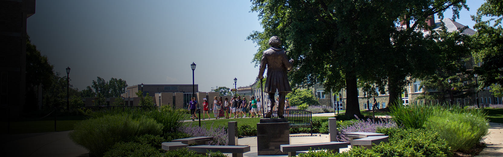 Statue of a historical figure standing on a pedestal in a landscaped campus courtyard, with students walking along a path in the background on a sunny day.