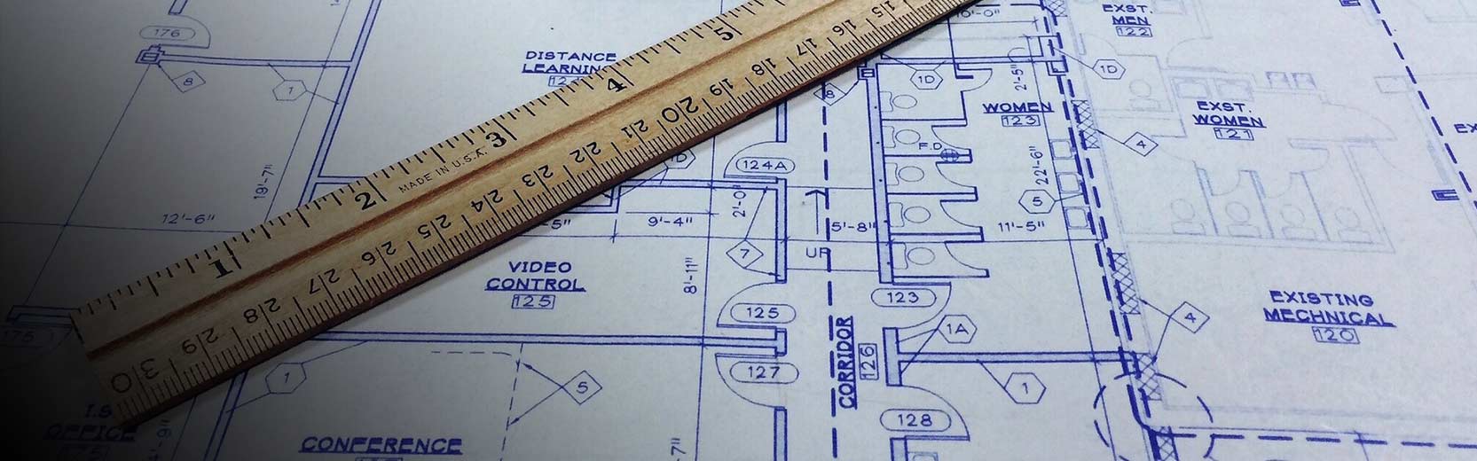 A close-up image of architectural blueprints with a wooden ruler placed on top. The blueprints show floor plans of various rooms, including labels for 