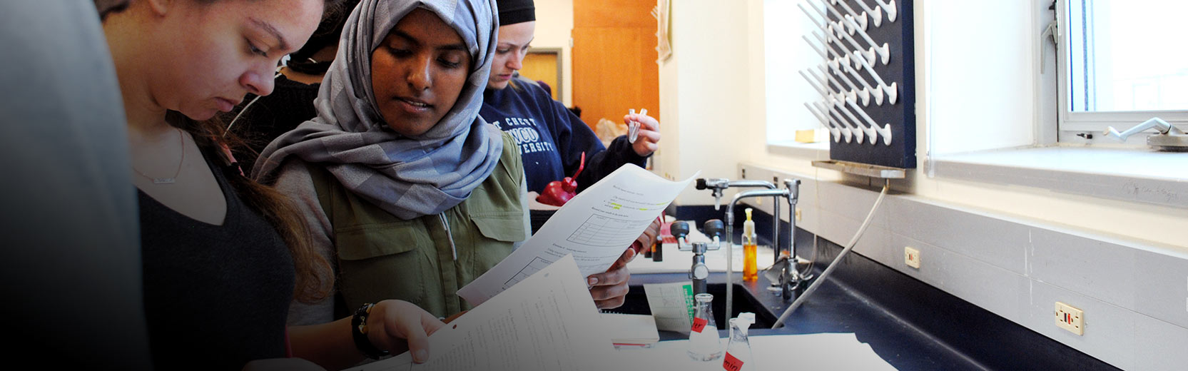 Students in a science lab review worksheets together at a counter with lab equipment, including flasks and a sink, while preparing for an experiment.