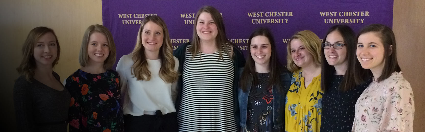 A group of eight women in front of a purple “West Chester University” backdrop, posing for a photo indoors.