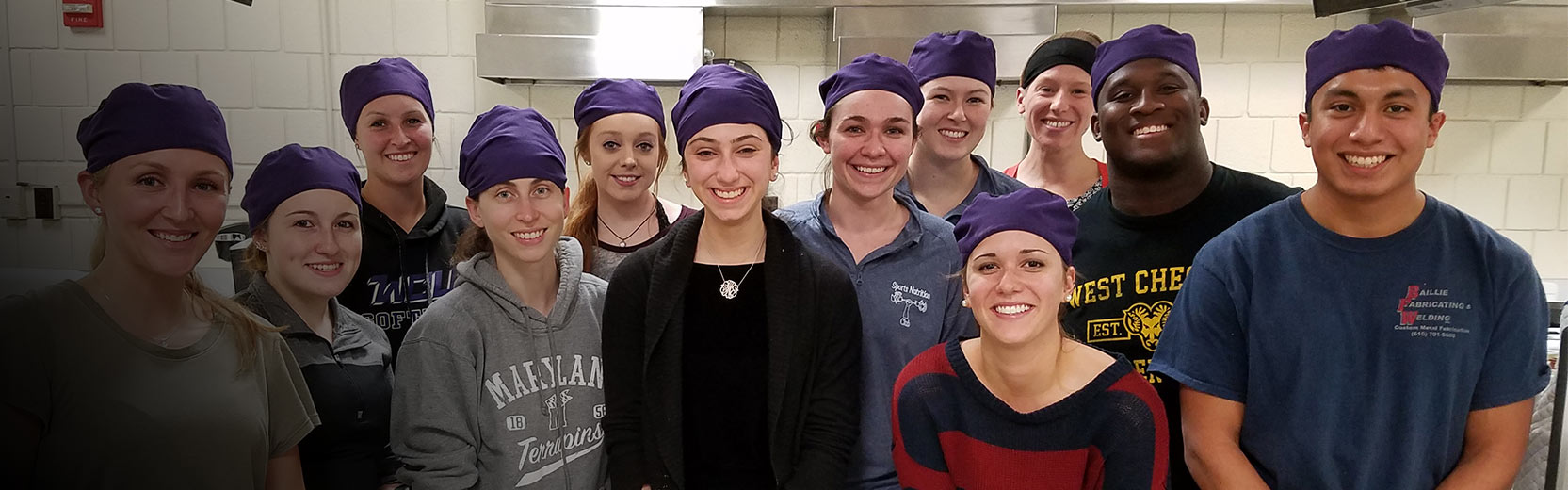 A group of smiling people wearing purple head coverings stand together in a kitchen or food preparation area, posing for a group photo.