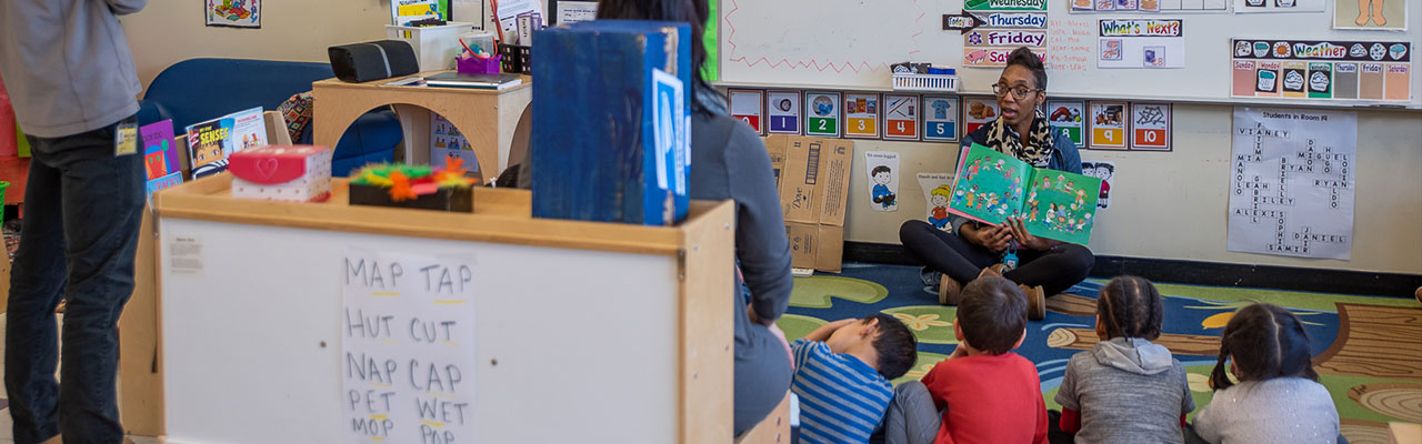 Person reading a book to 4 students that are sitting on the floor.